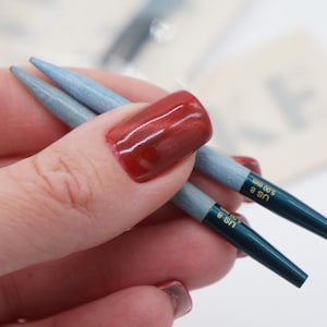 May include: Two blue knitting needles with gold lettering that reads "US 8 5.00 mm" and "US 6 4.00 mm".