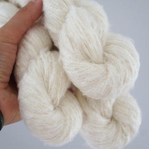 May include: Close-up of several skeins of soft, white yarn. The yarn appears fluffy and textured, ideal for knitting or crocheting projects. The yarn is held in a hand against a light grey background.