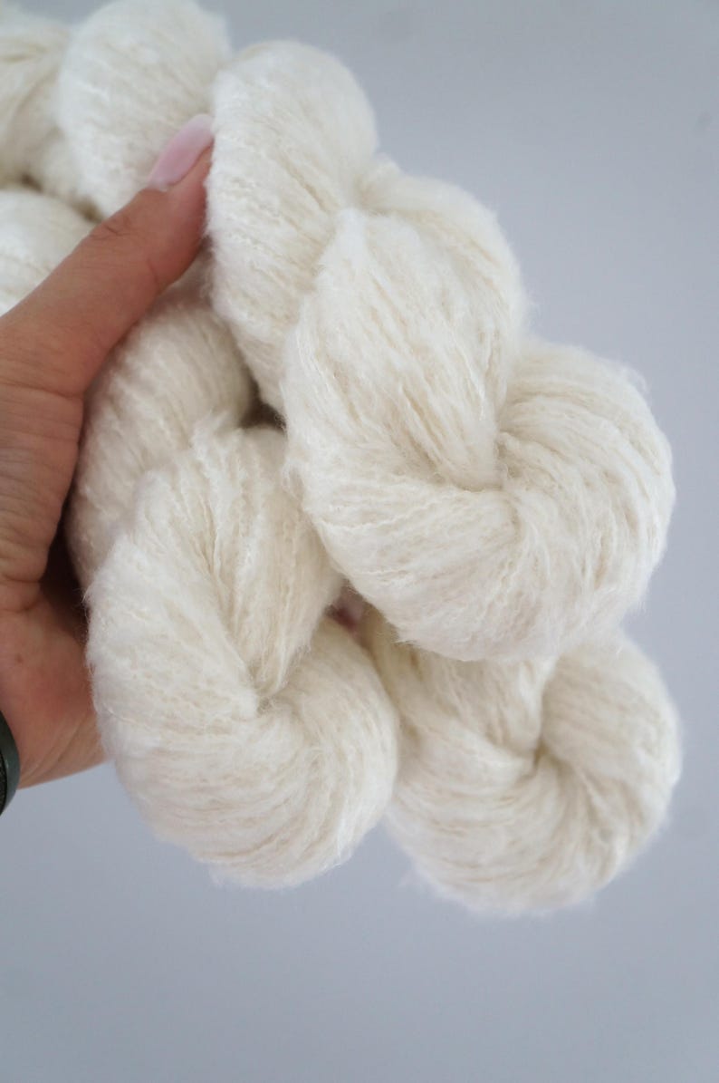 May include: Close-up of several skeins of soft, white yarn. The yarn appears fluffy and textured, ideal for knitting or crocheting projects. The yarn is held in a hand against a light grey background.