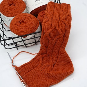 May include: A close-up of a knitted sock in a rust orange color with a cable knit pattern. The sock is being worked on with two knitting needles. Three balls of rust orange yarn are in a black wire basket. The yarn labels read "Ros&aacute;rios 4 Meia Classic Collection 50g 170m".
