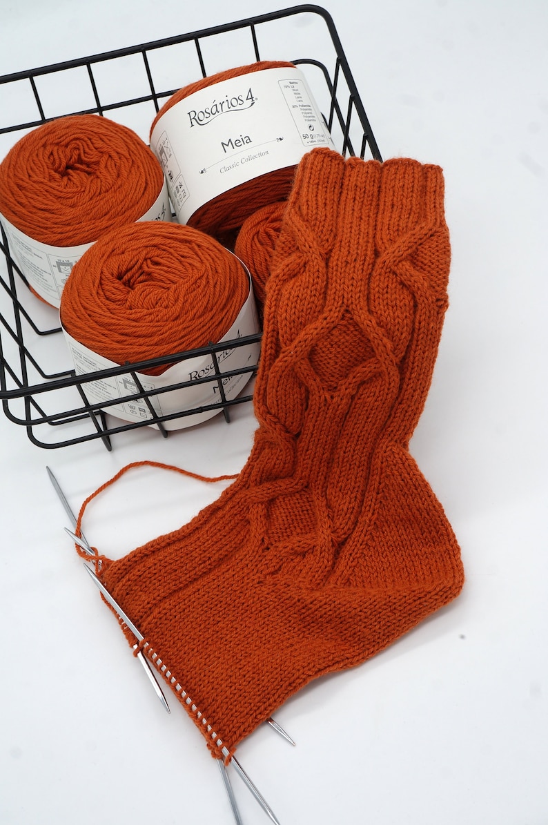 May include: A close-up of a knitted sock in a rust orange color with a cable knit pattern. The sock is being worked on with two knitting needles. Three balls of rust orange yarn are in a black wire basket. The yarn labels read "Ros&aacute;rios 4 Meia Classic Collection 50g 170m".