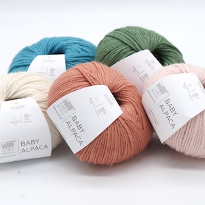 以下が含まれることがあります： 白、ターコイズブルー、緑、ピーチ、ピンクの5色の糸のボール。各ボールには、「ベビーアルパカ」と「カロウール」と書かれたラベルが付いています。