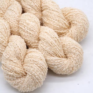 May include: Close-up of several skeins of cream-colored yarn. The yarn has a textured, nubby appearance, and is arranged in a braided pattern. The background is plain white, highlighting the yarn's natural color and texture.
