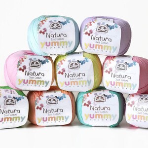 K&ouml;nnte beinhalten: Neun Kn&auml;uel DMC Natura Just Cotton Yummy Colors Garn in verschiedenen Pastellfarben, darunter Rosa, Gr&uuml;n, Blau und Gelb.