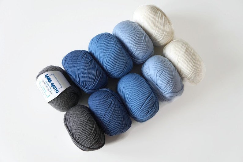 Puede incluir: Una colecci&oacute;n de ocho ovillos de lana en tonos de azul, gris y blanco. Los ovillos de lana est&aacute;n dispuestos en degradado de gris oscuro a blanco. La lana est&aacute; etiquetada como "Lana Gatto" y "Super Soft".