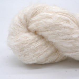 May include: A close-up shot of a skein of off-white yarn. The yarn has a soft, fluffy appearance with a slightly textured surface. The fibres are tightly twisted together, forming a dense bundle. The neutral colour suggests it's suitable for various knitting or crochet projects.
