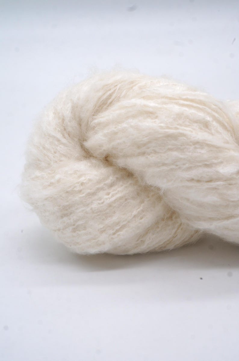 May include: A close-up shot of a skein of off-white yarn. The yarn has a soft, fluffy appearance with a slightly textured surface. The fibres are tightly twisted together, forming a dense bundle. The neutral colour suggests it's suitable for various knitting or crochet projects.
