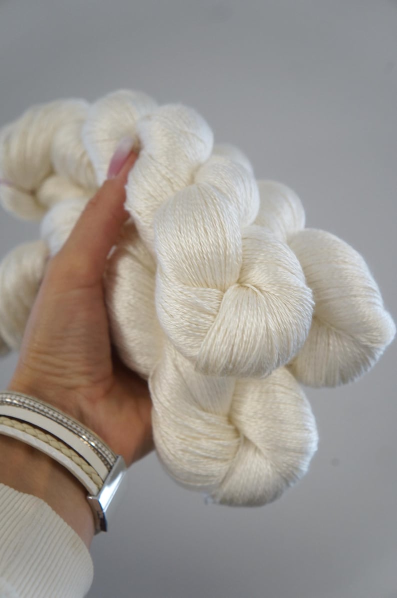 May include: A close-up of several skeins of off-white yarn. The yarn is held in a hand, showing the soft texture and the natural color. The yarn appears to be a natural fiber, perfect for knitting or crocheting projects.