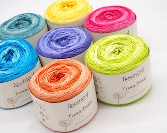 May include: A collection of colorful yarn skeins in shades of blue, green, yellow, pink, orange, and purple. Each skein is neatly wound and presented in a cylindrical paper wrapper. The yarn is labeled "Rosários 4 Troca-Tintas".