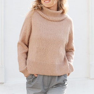 May include: A woman wearing a light brown turtleneck sweater with a ribbed texture. The sweater has a loose fit and a slightly cropped length. The woman is standing in front of a white wall. The image also includes the text "DROPS DESIGN" and "www.garnstudio.com".