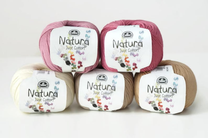 May include: Six balls of yarn in various shades of pink, beige, and cream. Each ball has a label that reads "Natura Just Cotton" with the DMC logo.