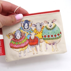 Op de afbeelding: Een crèmekleurig tasje met rits voorzien van een kleurrijke illustratie van vijf schapen die truien met patronen dragen. Een rood stoffen label aan de zijkant toont de tekst Emma Ball in het wit. Het tasje heeft een rode ritssluiting.