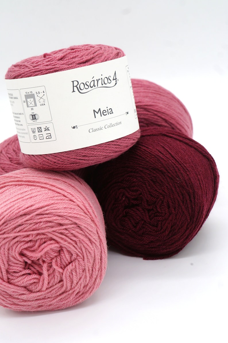May include: Three balls of yarn in shades of pink and burgundy. The label on the top ball reads "Ros&aacute;rios 4 Meia Classic Collection".