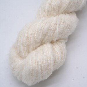 May include: Close-up of a skein of cream-coloured yarn. The yarn is thick and fluffy, with a soft, textured appearance. The fibres are loosely spun, creating a delicate, airy look. Suitable for knitting or crochet projects.