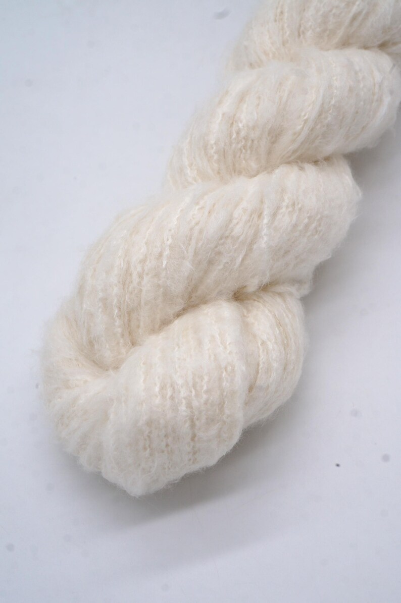 May include: Close-up of a skein of cream-coloured yarn. The yarn is thick and fluffy, with a soft, textured appearance. The fibres are loosely spun, creating a delicate, airy look. Suitable for knitting or crochet projects.