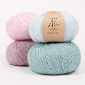 Puede incluir: Tres ovillos de lana en diferentes colores: rosa, morado y azul. El ovillo azul tiene una etiqueta que dice "DROPS Air uni colour" y "Find free knitting & crochet patterns at garnstudio.com".