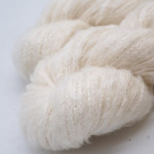 May include: Close-up of a skein of off-white yarn. The yarn appears soft and fluffy, with a slightly textured surface. The fibres are tightly spun together, creating a dense, yet airy appearance. The yarn is ideal for knitting or crocheting projects.