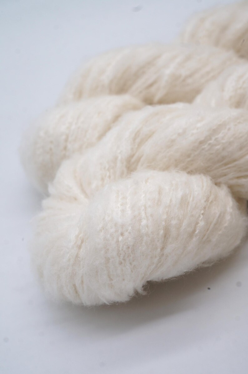 May include: Close-up of a skein of off-white yarn. The yarn appears soft and fluffy, with a slightly textured surface. The fibres are tightly spun together, creating a dense, yet airy appearance. The yarn is ideal for knitting or crocheting projects.