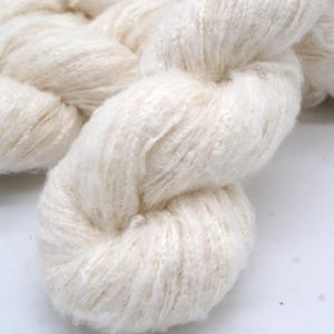 May include: Close-up of a thick, off-white yarn braid. The yarn appears soft and fluffy, with a slightly textured surface. The fibers are tightly twisted together, creating a dense, rope-like structure. Ideal for knitting or crocheting projects.