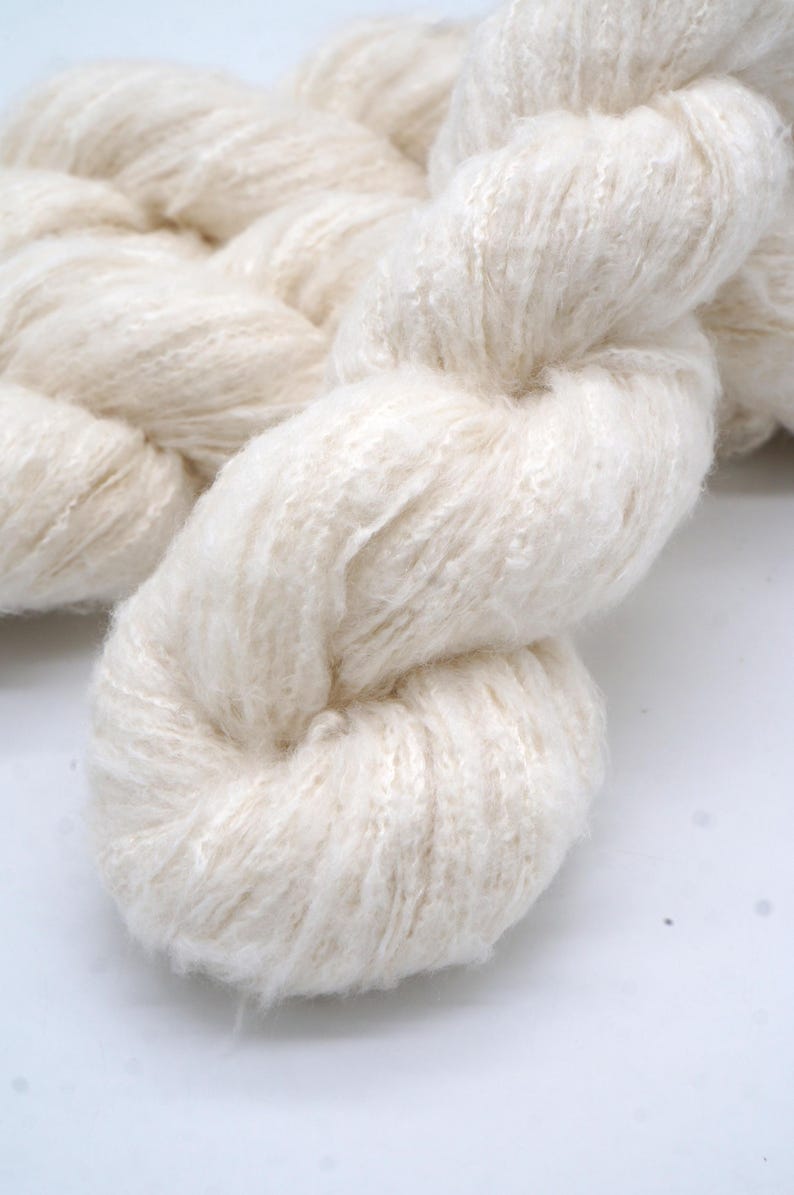 May include: Close-up of a thick, off-white yarn braid. The yarn appears soft and fluffy, with a slightly textured surface. The fibers are tightly twisted together, creating a dense, rope-like structure. Ideal for knitting or crocheting projects.