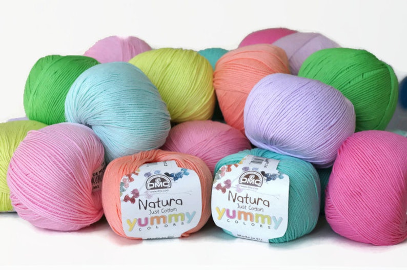 K&ouml;nnte beinhalten: Eine Sammlung bunter Wollkn&auml;uel in den Farben Rosa, Gr&uuml;n, Blau und Orange. Das Garn ist mit "Natura Just Cotton Yummy Colors" gekennzeichnet und wird von DMC hergestellt.