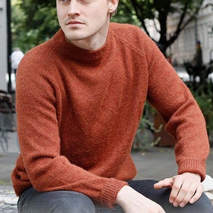 May include: A rust-colored sweater with a round neckline and long sleeves. The sweater is made of a soft, fuzzy material. The text "DROPS DESIGN" and "www.garnstudio.com" is visible at the bottom of the image.