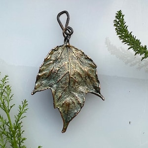 May include: A metal pendant shaped like a maple leaf with a textured, oxidized finish in shades of bronze, copper, and verdigris green. The leaf features a prominent central vein and jagged edges, attached to a dark, twisted metal wire loop bail.