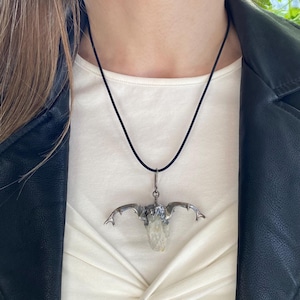 May include: A necklace featuring a black cord and a silver-toned metal pendant shaped like a skull with branching antlers. The center of the pendant holds a translucent, pale yellow crystal stone set within the metallic structure.