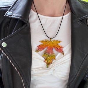 May include: A maple leaf pendant necklace with a black cord. The leaf is a mix of orange, yellow, and red. The necklace is worn over a white shirt and black leather jacket.