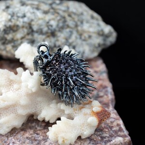 May include: A dark gunmetal gray pendant shaped like a sea urchin, featuring a dense cluster of sharp, radiating spikes and a small circular bail at the top for attachment. The metallic surface has a polished finish that reflects light.