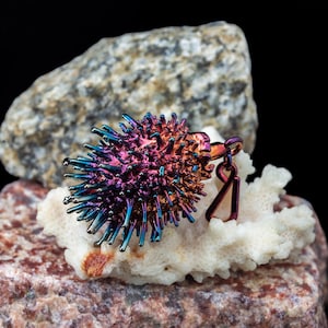 May include: A multi-coloured, spiky metal pendant, resembling a sea urchin, with shades of purple, blue, and orange. The pendant is displayed on white coral and a textured rock, set against a black backdrop. The pendant has a small metal loop.