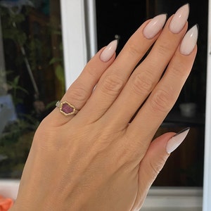 May include: A hand wearing a silver ring with a gold bezel holding a rough-cut, pink gemstone. The nails are painted with a light pink base and black tips, creating a French manicure style.