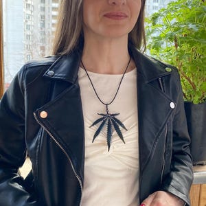 May include: A black cannabis leaf pendant suspended on a thin black cord necklace. The pendant features a detailed, serrated leaf design with a dark, matte finish. It is worn over a plain white top and a black leather jacket with silver zippers.