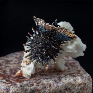 May include: A dark, metallic, spherical pendant featuring numerous protruding spikes with gold-toned tips. The pendant has a textured, oxidized finish and is attached to a thin, dark metal chain loop at the top, showcasing a complex, organic form.