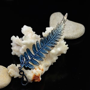 May include: A metallic fern leaf pendant featuring a gradient of deep blue and dark grey tones. The leaf has a detailed, segmented structure with a small circular loop at the base for attachment, showcasing a reflective, iridescent finish.