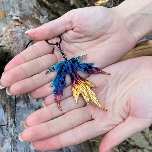 May include: A pendant shaped like a maple leaf with a metallic finish that transitions in color from deep blue and purple at the top to vibrant orange and gold at the bottom, attached to a thin black cord necklace.