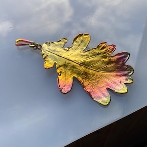 Può includere: Un ciondolo metallico a forma di foglia di quercia con una finitura sfumata che passa dal giallo e oro brillante a tonalità magenta e viola intenso. La superficie presenta venature in rilievo dettagliate e un bordo strutturato, fissato a un occhiello.