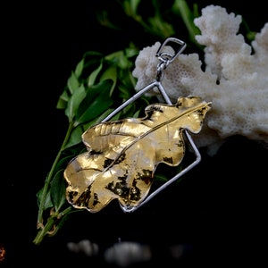 Op de afbeelding: Een gouden eikenblad hanger met een zilveren frame. De hanger heeft gedetailleerde bladnerven. De achtergrond is zwart, met groene bladeren en een wit, koraalachtig object.