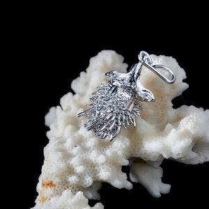 May include: A silver-toned pendant shaped like a spiky, textured seed pod or burr. The metallic piece features a small loop at the top for attachment to a chain. The surface is highly reflective with numerous thin, protruding spikes.