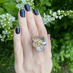 May include: A statement ring featuring a large, dimensional flower design. The petals have a textured, silver-toned finish with gold-toned edges, while the centre of the flower is a raised, textured gold-toned disc. The ring is worn on a finger.