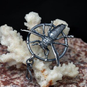 May include: A dark metallic pendant featuring a detailed insect, likely a wasp or bee, positioned within a circular frame. The pendant has a textured, oxidised finish and includes a small loop at the bottom for attachment to a chain or cord.