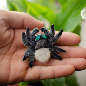 May include: A small, gray and white spider with blue eyes. The spider is made of felt and has a fuzzy texture.