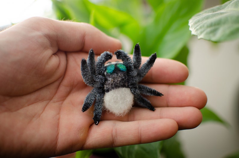 May include: A small, gray and white spider with blue eyes. The spider is made of felt and has a fuzzy texture.