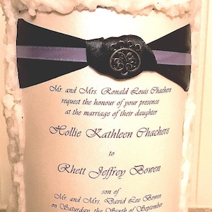 May include: A large, white candle with a textured, melted wax appearance. A black ribbon with a silver stripe and a decorative emblem adorns the candle. The candle features printed text announcing a wedding.