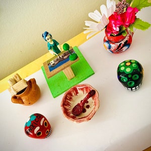 May include: A collection of colorful, handcrafted Mexican folk art items. Includes a small ceramic pot with flowers, painted skulls, a miniature sign, a small bowl with a spoon, and a clay pot with sticks.
