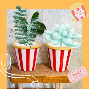 May include: Two decorative ceramic planters with red and white stripes, reminiscent of popcorn buckets. One planter holds greenery and small flowers, the other a light green succulent. The planters sit on a small wooden shelf.