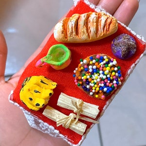 May include: Miniature food items on a red fabric square with white lace trim. Includes a loaf of bread, a green and pink object, a donut with sprinkles, a purple ball, a banana, and two tied bundles.