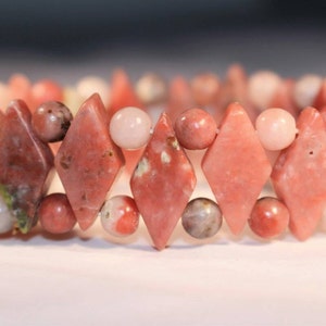 May include: A beaded bracelet featuring a mix of pink and red gemstones. The bracelet has diamond-shaped and round beads in varying shades of pink and red. The bracelet is a beautiful piece of jewelry.