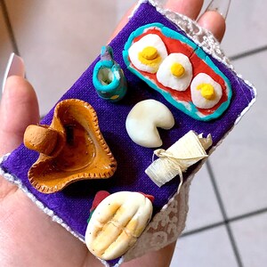 May include: Miniature food items arranged on a purple square with a white lace trim. The set includes a plate of eggs, a small cup, a bread roll, a mushroom, and a small container. The food items are brightly colored and detailed.