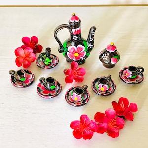 May include: Miniature black ceramic tea set with pink floral designs, including a teapot, sugar bowl, and several teacups with saucers. Bright pink flowers are scattered around the set, creating a charming and decorative display.